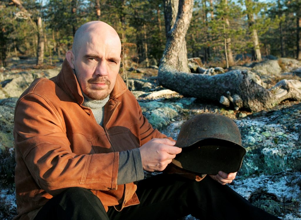 Pekka Jaatinen istuu maastossa sylissään sota-aikainen kypärä.